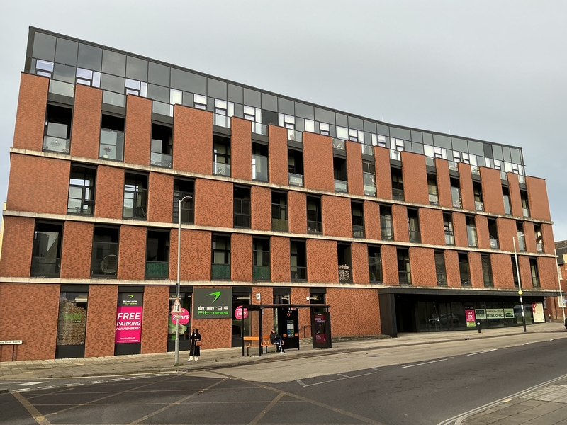 5 story commercial building and flats / apartments. Bus stop on the road. Grey sky.