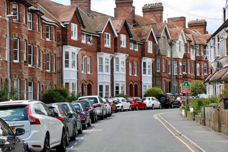Row of 3 story town houses, red brick. cars parked along side the road. one of the houses has a for sale sign outside.
